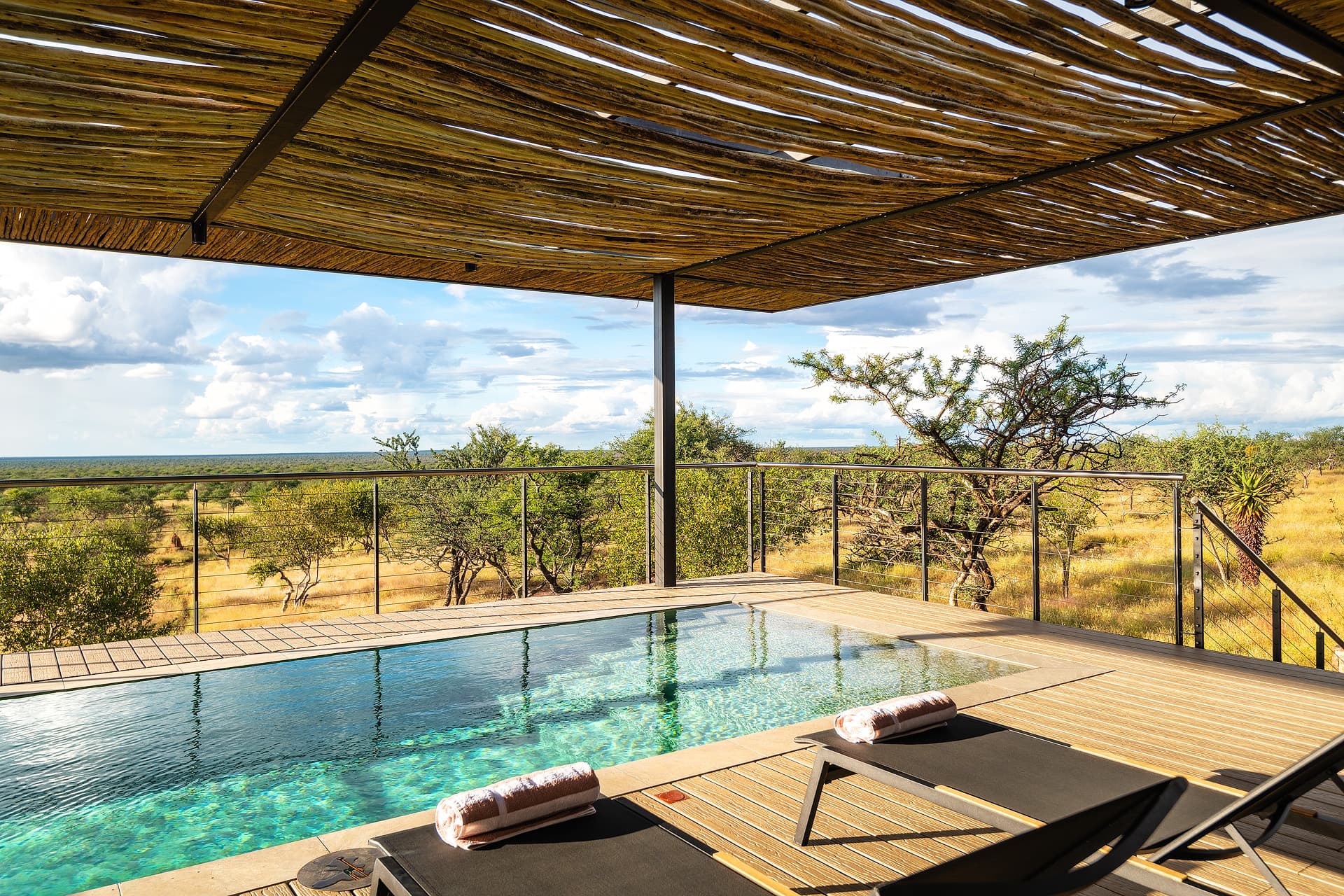 Luxury pool deck with lounge chairs overlooking a vast savanna under a rustic wooden roof.