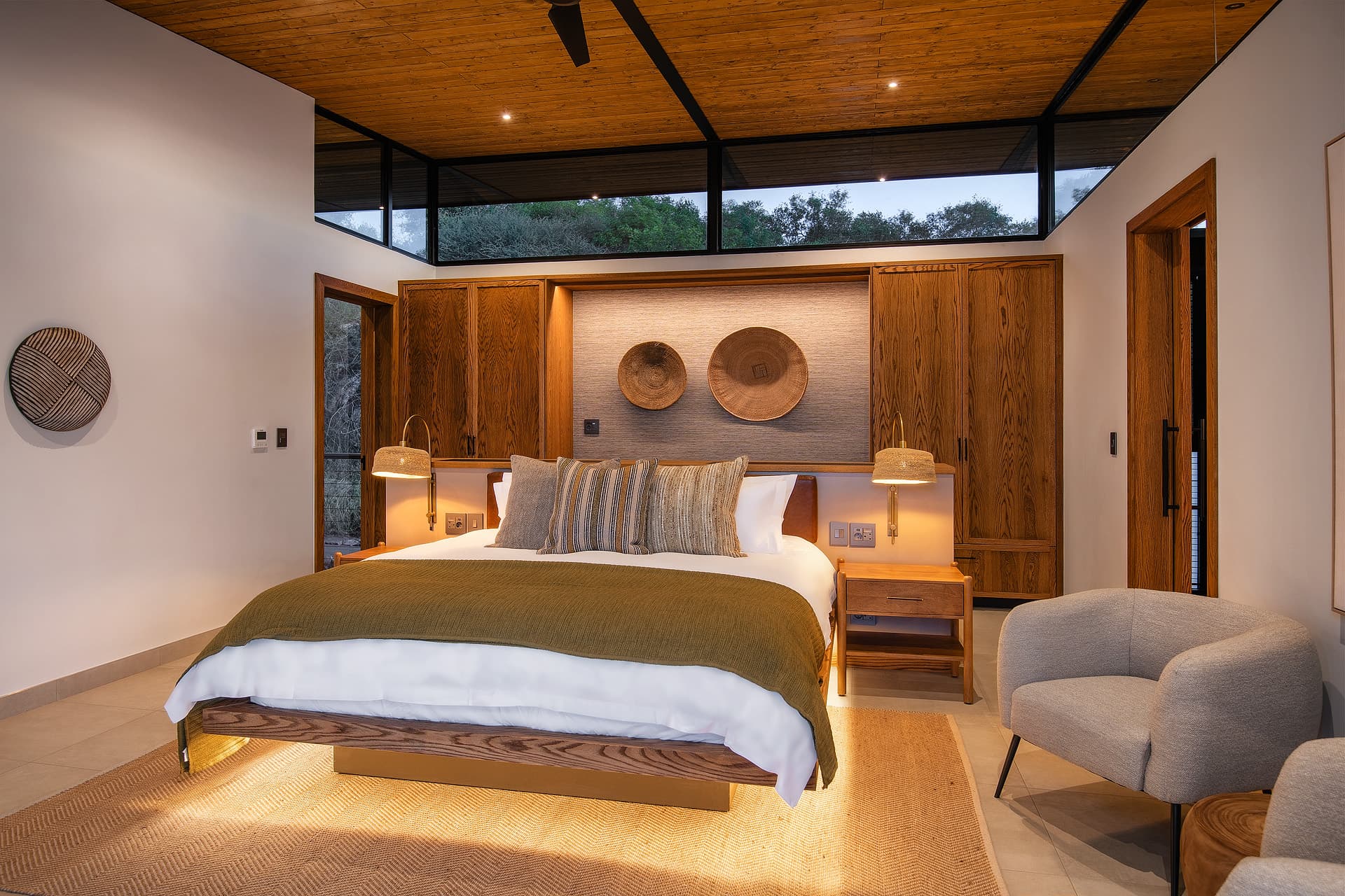 Modern bedroom with wooden ceiling, olive green bedding, warm lighting, and woven wall decor.
