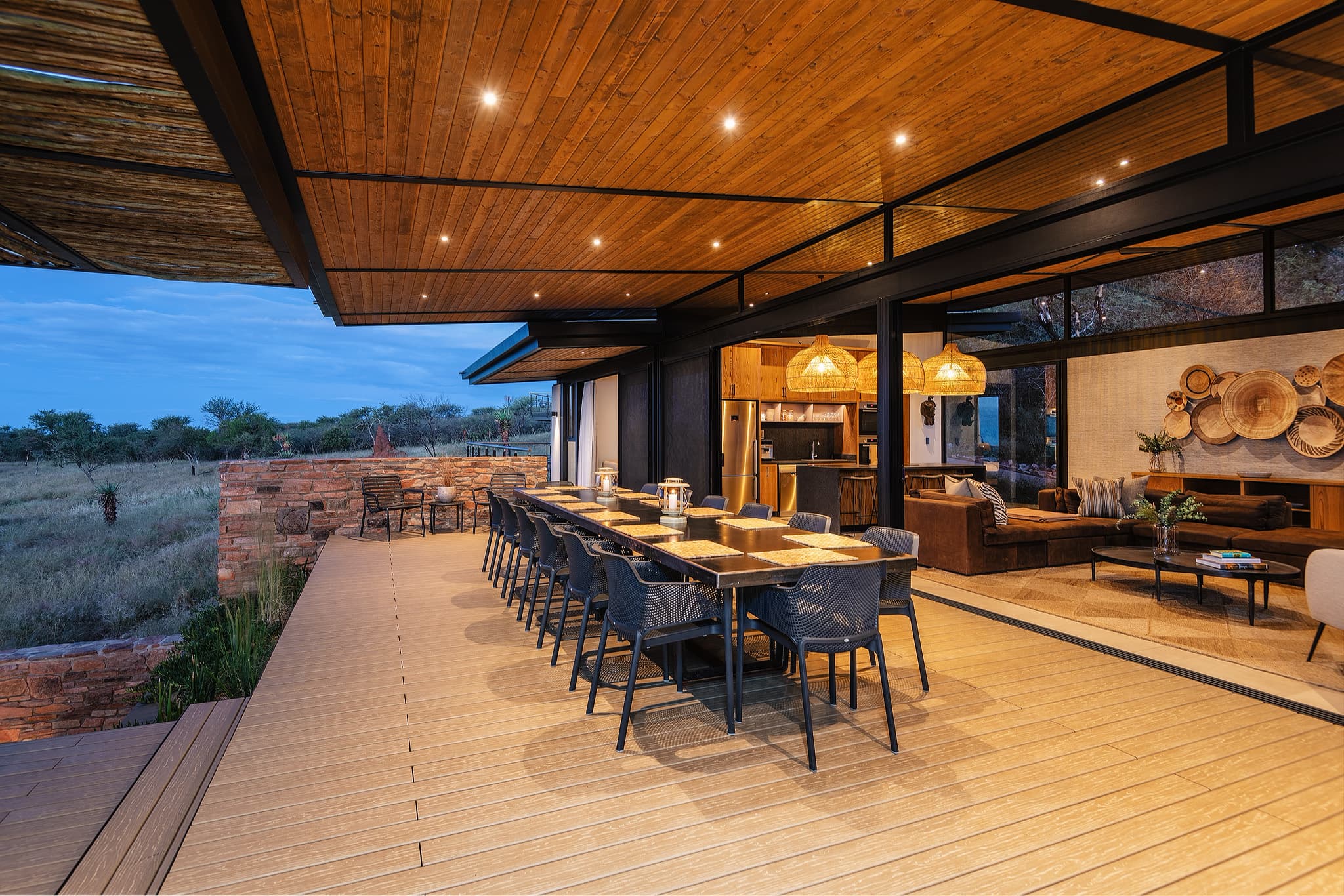Luxurious wooden deck with long dining table and warm lighting overlooking a savanna at dusk.