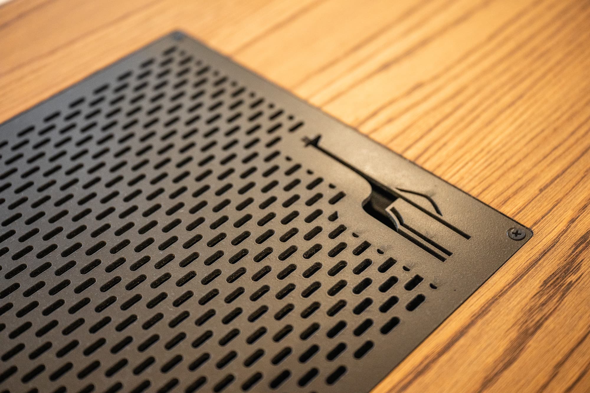Black perforated metal grate with a latch mechanism set into a light-colored wooden surface.
