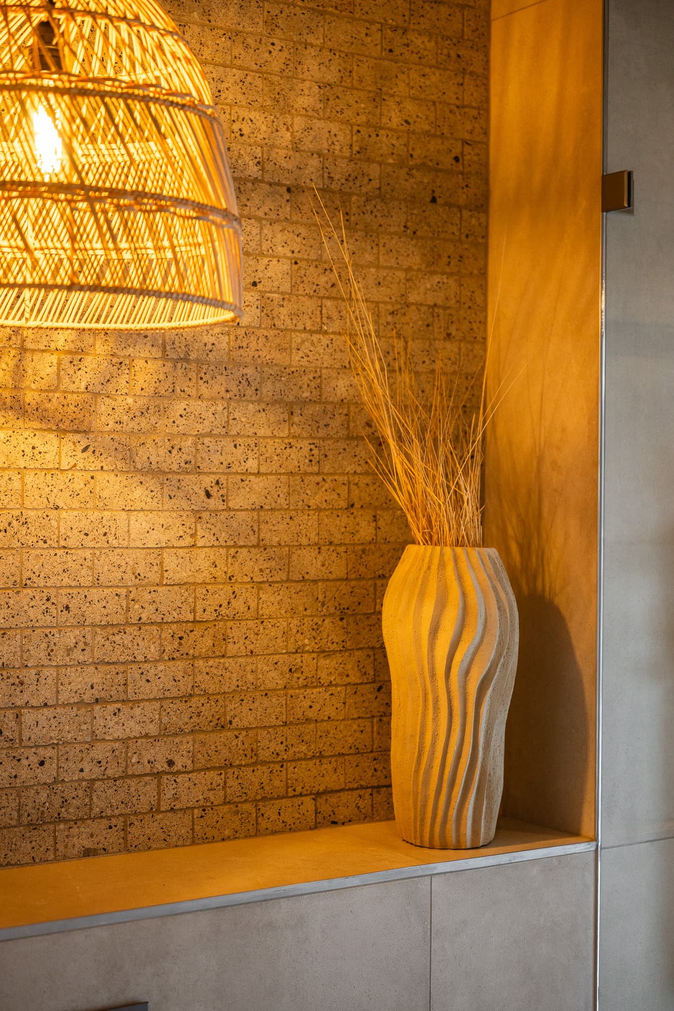 Tall wavy vase with dried grass under warm light from a wicker pendant lamp.