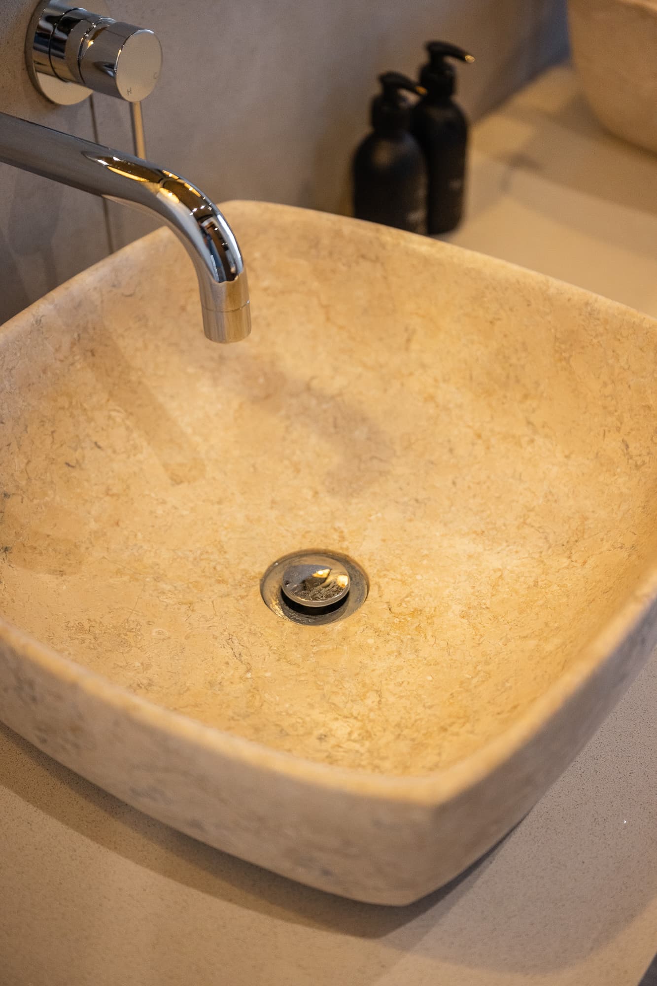 Square beige stone vessel sink with a wall-mounted chrome faucet and black soap dispensers.