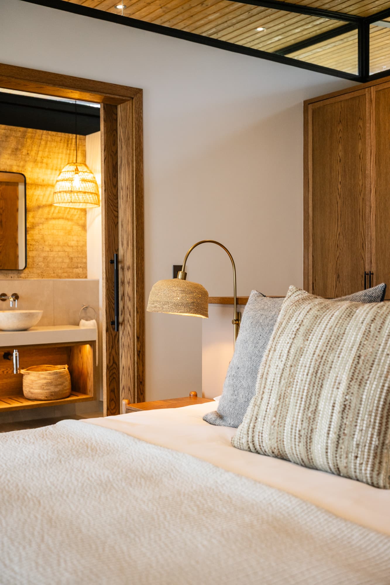 Modern bedroom with neutral textured pillows, brass woven lamp, and view into a stone-tiled bathroom.